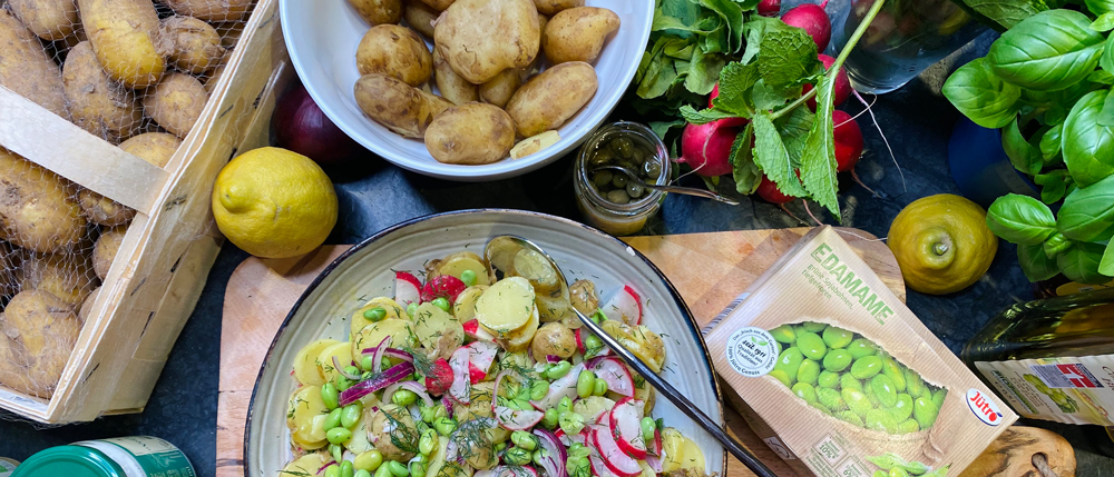 Schüssel mit Kartoffelsalat, Edamame und Radieschen, daneben rohe Zutaten wie Kartoffeln, Zitronen und frische Kräuter.