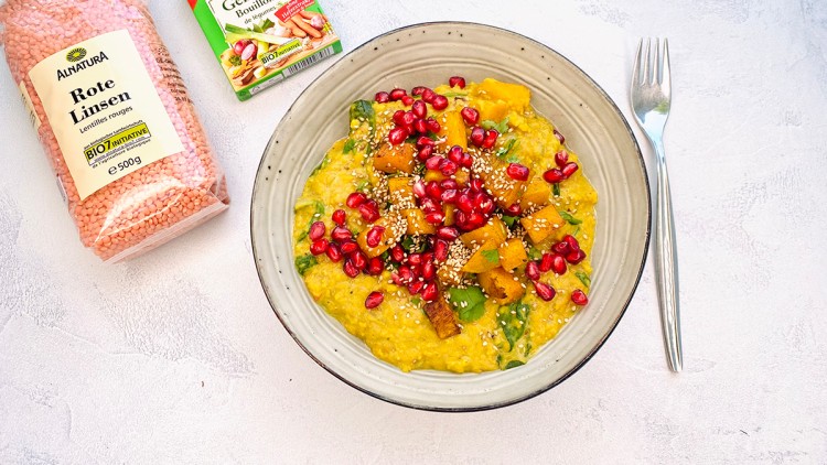 Graue Schale mit Linsengericht, Kürbiswürfeln, Granatapfelkernen und Sesam neben roter Linsenpackung, Gemüsebouillon und Gabel.