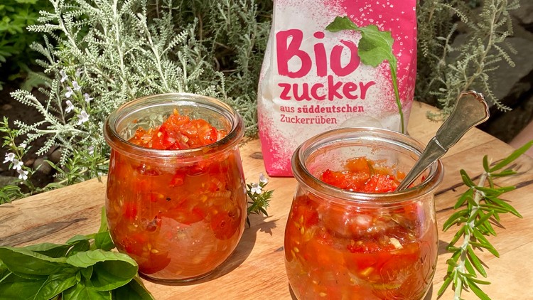 Zwei Gläser mit Tomatenchutney, frische Kräuter und eine Packung Biozucker aus süddeutschen Zuckerrüben auf Holztisch.