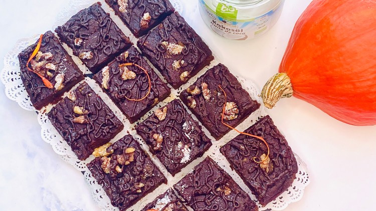 Zwölf Schokoladen-Brownies mit Walnüssen und Kürbisdekoration auf Spitze, daneben ein Hokkaidokürbis und ein Glas Kokosöl.