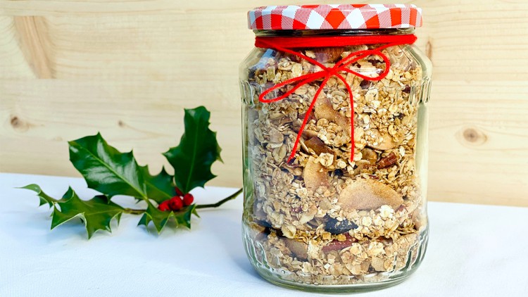 Ein Glas mit Müsli, verziert mit rotem Band und kariertem Deckel, steht neben einem Zweig mit roten Beeren.