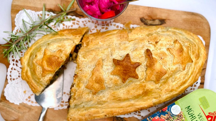 Ein goldbrauner Blätterteig mit Stern- und Tannenbaumverzierungen liegt auf Holzbrett, daneben bunt eingelegtes Gemüse in Glas.
