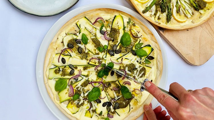 Hand schneidet eine Gemüse-Pizza mit Zucchini, roten Zwiebeln, Basilikum, Kürbiskernen und grünem Pesto auf weißem Teller.