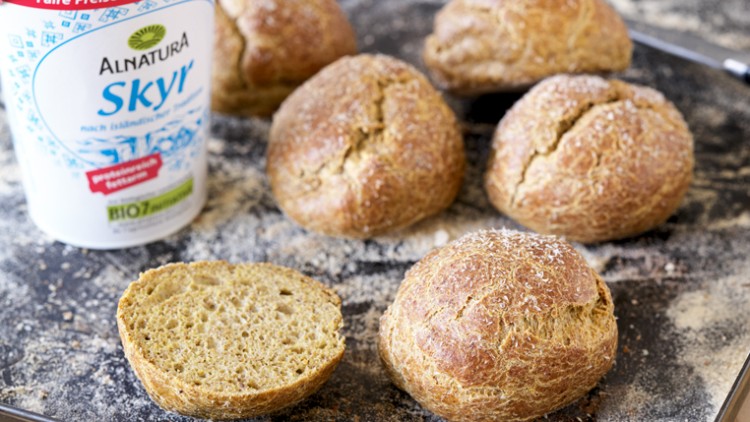 Fünf gebackene Brötchen und ein aufgeschnittenes Brötchen mit einer Packung „Alnatura Skyr“ auf bemehlter Fläche, daneben ein Messer.