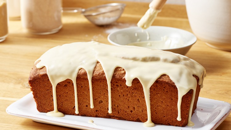 Rechteckiger Kuchen auf einer weißen Platte, mit hellem Zuckerguss überzogen, im Hintergrund Backzutaten und Schüssel.