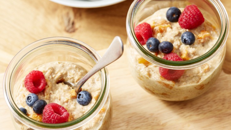 Zwei Gläser mit Haferbrei, garniert mit Himbeeren und Blaubeeren, stehen auf einem Holzbrett, ein Löffel steckt im vorderen Glas.