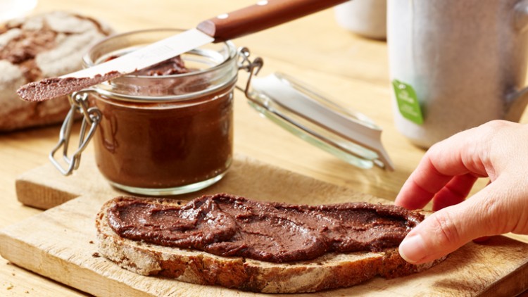 Eine Hand greift nach einer Scheibe Brot mit braunem Aufstrich auf einem Holzbrett, im Hintergrund ein Glas mit Aufstrich und eine Tasse.