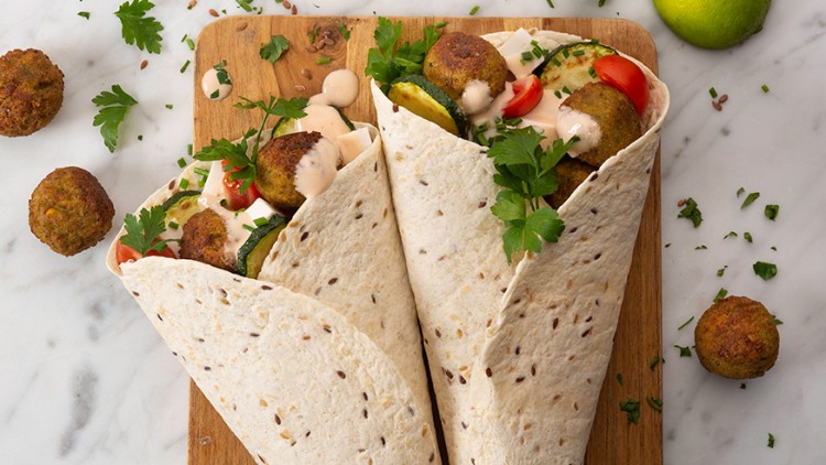 Zwei Wraps mit Falafel, Tomaten, Gurken, Petersilie und heller Soße auf einem Holzbrett, dazu Limetten und Kräuter auf Marmor.