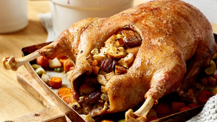 Eine goldbraun gebratene Gans, gefüllt mit Brot und Maronen, liegt auf einem Blech mit geröstetem Gemüse wie Karotten und Lauch.