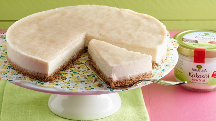 Auf einer Tortenplatte steht eine helle Torte mit einer herausgenommenen Scheibe, daneben ein Glas Alnatura Kokosöl neutral.
