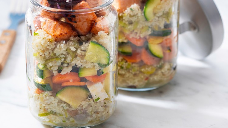 Zwei Einmachgläser mit Schichten aus Quinoa, Gemüse, angebratenem Lachs und purpurfarbenen Sprossen auf weißem Untergrund.