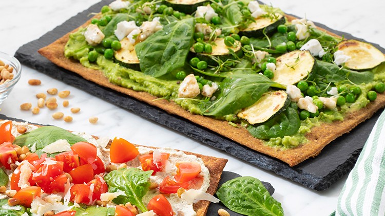 Zwei dünne Fladen mit Gemüse, Blattspinat, Crème, Tomaten und Zucchini auf schwarzen Schieferplatten, daneben Pinienkerne.