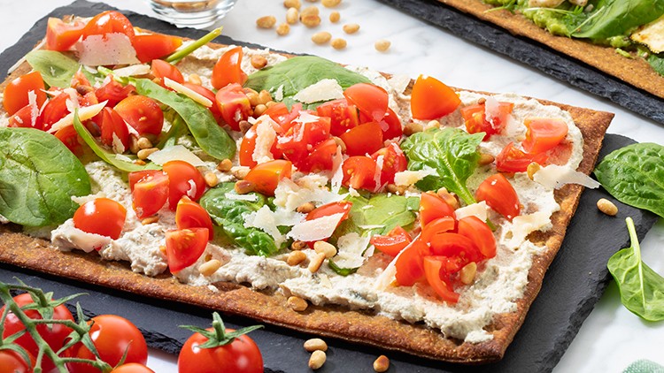 Zwei rechteckige Flammkuchen mit frischem Gemüse wie Spinat, Tomaten, Zucchini und Erbsen auf Schieferplatten, daneben Pinienkerne.