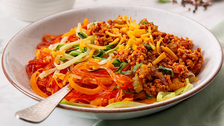 Weiße Schale mit Gemüsenudeln, vegetarischer Hackfleischsauce und geriebenem Käse auf grünem Stofftuch, im Hintergrund Gewürze.