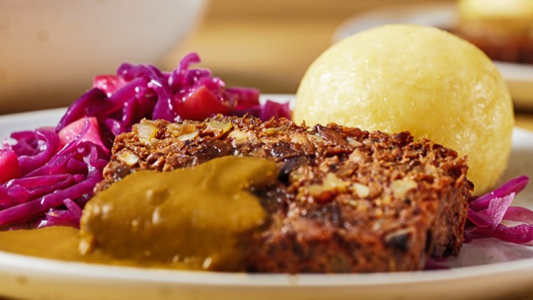 Nahaufnahme von veganem Braten mit brauner Sauce, rotem Krautsalat und einem Kartoffelknödel auf einem weißen Teller.