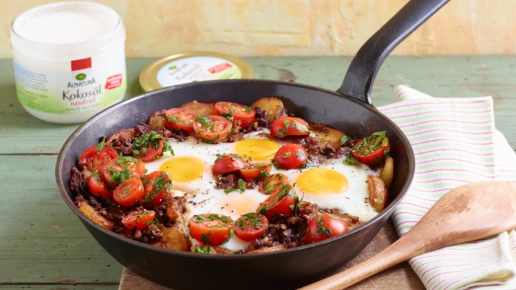 Eine schwarze Pfanne mit Spiegeleiern, Tomaten, Kräutern und Kartoffeln steht auf Holztisch mit Holzlöffel und Kokosöl im Hintergrund.