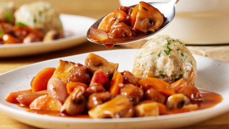 Ein weißer Teller mit Pilzragout, Karotten und einem Serviettenknödel, eine Löffelportion wird darüber gehalten.