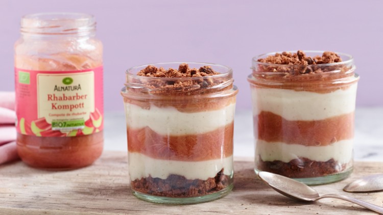 Zwei Gläser mit Schichten aus Rhabarberkompott, Creme und Streuseln stehen auf einem Holzbrett, daneben ein Kompottglas.