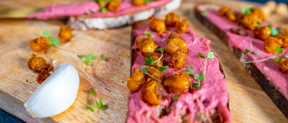 Dunkles Brot mit pinkem Aufstrich, gerösteten Kichererbsen, Microgreens und einer Knoblauchzehe auf einem Holzbrett.