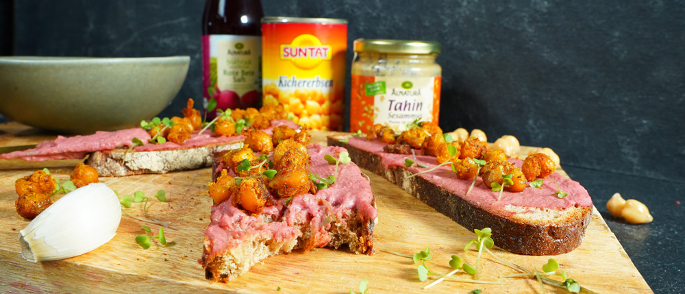 Drei Brotscheiben mit rosa Aufstrich, gerösteten Kichererbsen und Sprossen auf Holzbrett, dahinter Zutaten wie Tahin und Kirschsaft.