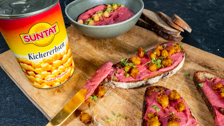 Eine geöffnete SUNTAT Kichererbsen-Dose neben Brot mit rosa Aufstrich, gebratenen Kichererbsen und Kräutern auf einem Holzbrett.