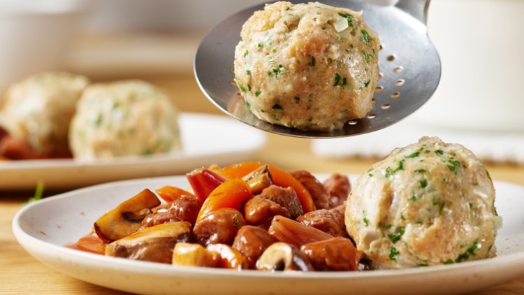 Auf einem Teller liegen zwei Semmelknödel mit Petersilie und ein Ragout aus Champignons, Paprika und Möhren, weiteres im Hintergrund.