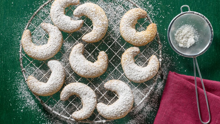 Halbmondförmige Plätzchen mit Puderzucker auf einem Gitter, daneben ein Sieb mit Puderzucker und ein violettes Tuch.
