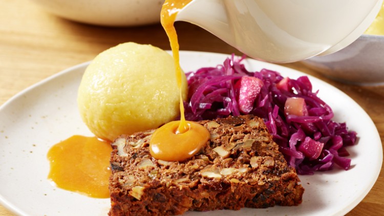 Ein Teller mit veganem Braten, rotem Krautsalat und Kartoffelklößen, übergossen mit heller Soße aus einer weißen Kanne.