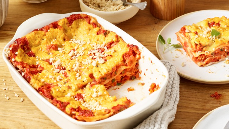 Eine Auflaufform mit gebackener Lasagne, daneben ein Teller mit einem servierten Stück und Besteck auf einem Holztisch.