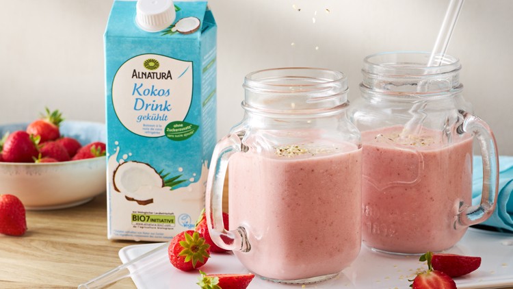 Zwei Gläser mit rosa Erdbeer-Smoothie neben einer Packung Alnatura Kokos Drink und einer Schale mit frischen Erdbeeren.