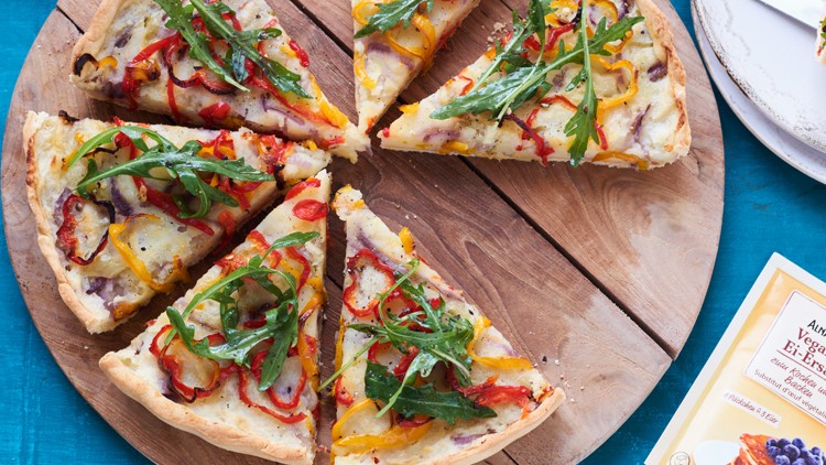 Eine runde Gemüsepizza mit Rucola, in Stücke geschnitten, liegt auf einem Holzbrett, daneben Alnatura Veganer Ei-Ersatz.