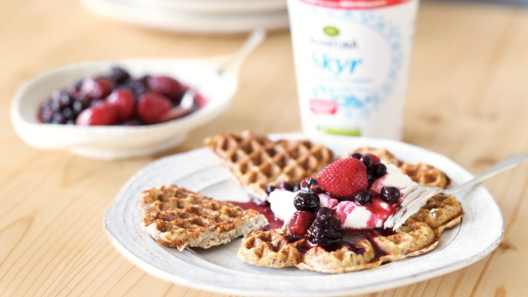Vollkornwaffeln mit Joghurt und roten Beeren auf einem Teller, daneben Schale mit Beeren und Becher „Skyr“ auf Holztisch.