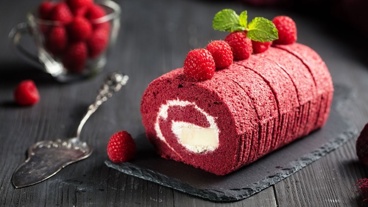 Rote Biskuitrolle mit heller Cremefüllung, dekoriert mit Himbeeren und Minzblättern, auf einer dunklen Schieferplatte.