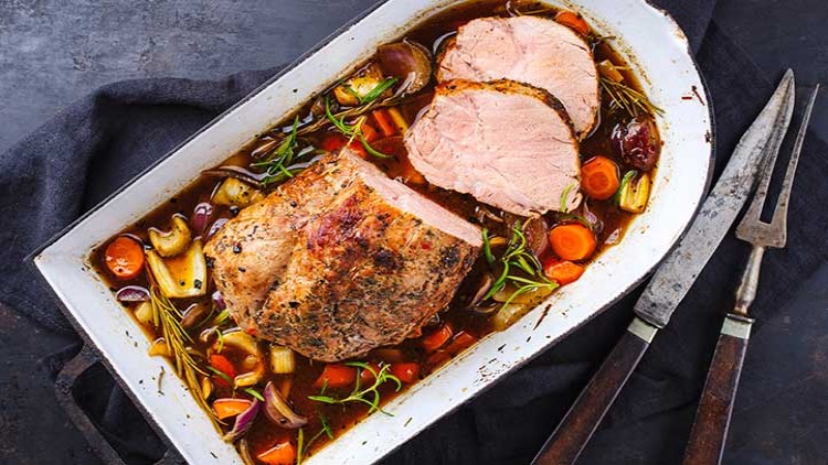 Ein Braten mit Kräutern, Zwiebeln, Karotten und Soße liegt in einer weißen Auflaufform neben Besteck auf dunklem Untergrund.