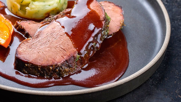 Zwei Scheiben Braten mit dunkler Soße, Petersilienkartoffelpüree und einem Stück Karotte auf einem dunklen Teller.