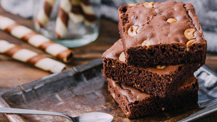 Drei rechteckige Schokoladenbrownies mit Nüssen gestapelt auf einem Tablett, im Hintergrund Glas mit braun-weißen Gebäckstangen.