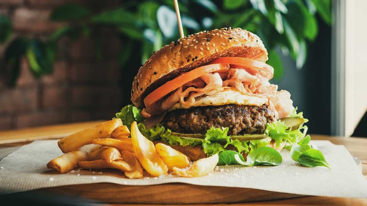 Hamburger mit Sesambrötchen, Salat, Tomaten, Fleischpatty, Ei und Schinken, dazu Pommes frites auf brauner Unterlage.