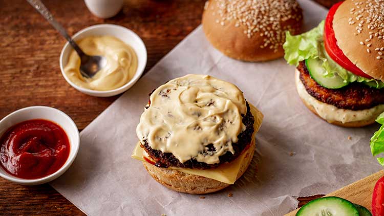 Ein Hamburgerbrötchen mit Fleischpatty, Käse und heller Soße, daneben Schüsseln mit Ketchup und Mayonnaise, Salat und Gemüse.