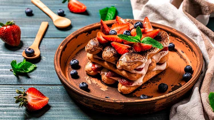 Ein Teller mit Tiramisu, dekoriert mit frischen Erdbeeren, Blaubeeren und Minzblättern, auf einem Holztisch mit Besteck.