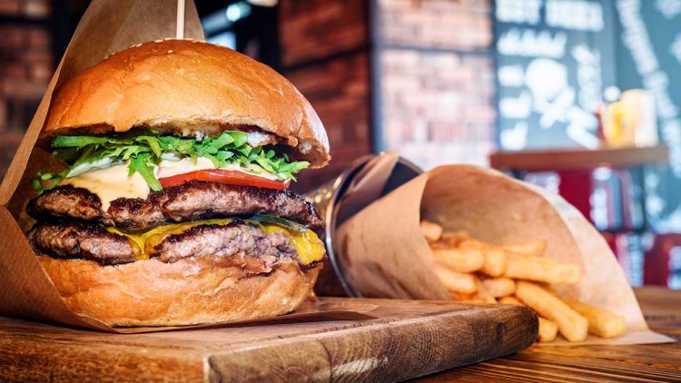 Ein Burger mit zwei Fleischpattys, Käse, Tomaten, Salat und Soße liegt auf einem Holzbrett, daneben Pommes in einer Papiertüte.