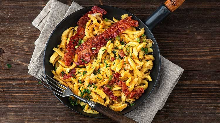 Schwarze Pfanne mit Käsespätzle, knusprigem Speck und gehackter Petersilie auf einem hellgrauen Stofftuch auf Holztisch.