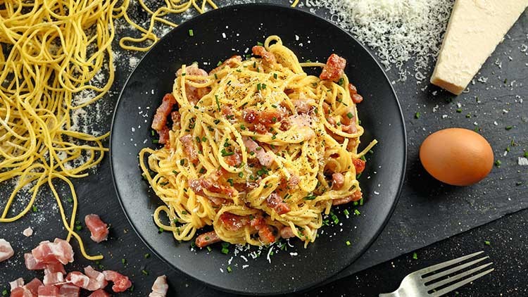 Ein Teller Spaghetti Carbonara mit Speck, Ei und geriebenem Käse, daneben rohe Nudeln, Parmesan, Speckwürfel und ein Ei.