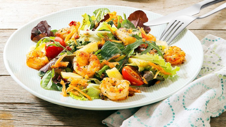 Ein Teller mit gemischtem Blattsalat, gebratenen Garnelen, geriebenen Karotten, Tomaten und Ananasstücken, daneben Besteck.