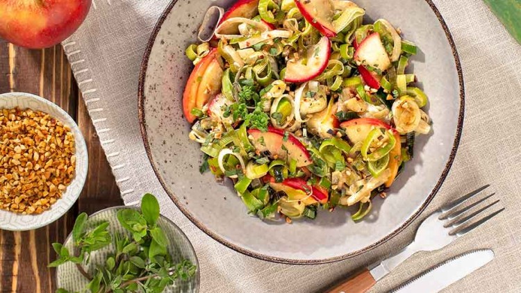 Teller mit Lauch-Apfelsalat, daneben Apfel, Lauch, Kräuter, Nüsse, Butter und Besteck auf Holzuntergrund mit Stoffserviette.
