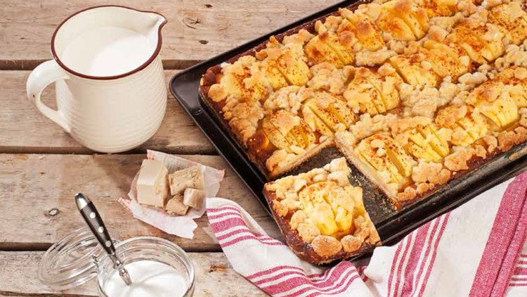 Blechkuchen mit Streuseln und Äpfeln auf Holztisch, daneben Milchkanne, Zucker im Glas, Butter, Hefe und rot-weißes Küchentuch.