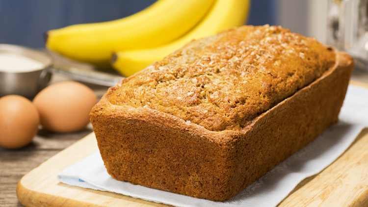 Ein Laib Bananenbrot auf einem Holzbrett, daneben zwei Bananen, zwei Eier und eine Schüssel mit Mehl im Hintergrund.