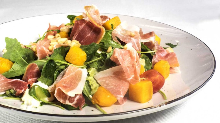 Salat mit Rucola, Parmaschinken, gelben Melonenwürfeln, Kirschtomaten, Pinienkernen und Dressing auf einem weißen Teller.