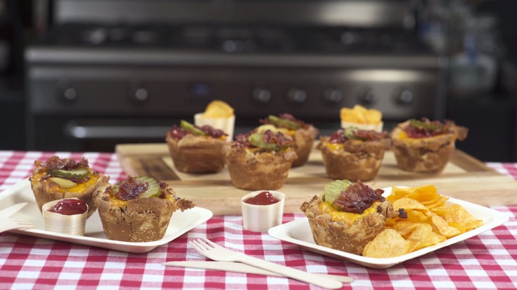 Auf einem Holztisch stehen Muffins mit Käse, Gurkenscheiben und Bacon, daneben Teller mit Chips und Ketchup auf karierter Tischdecke.