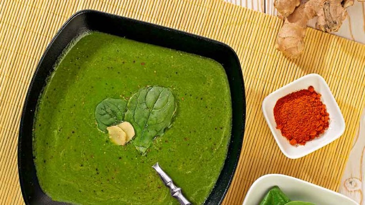 Grüne Suppe in schwarzer Schale mit Blattspinat und Ingwer garniert, daneben Limette, Ingwerwurzel, Gewürzpulver und frischer Spinat.