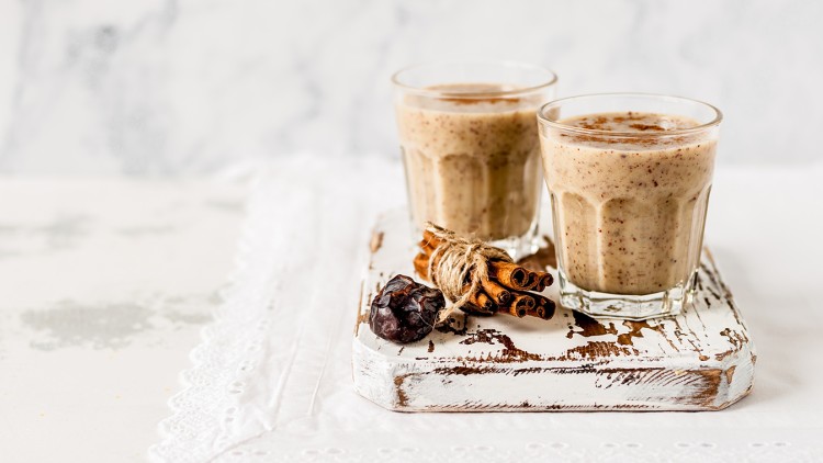Zwei Gläser mit cremigem Smoothie stehen auf einem weißen Holzbrett, daneben Zimtstangen und eine getrocknete Dattel.